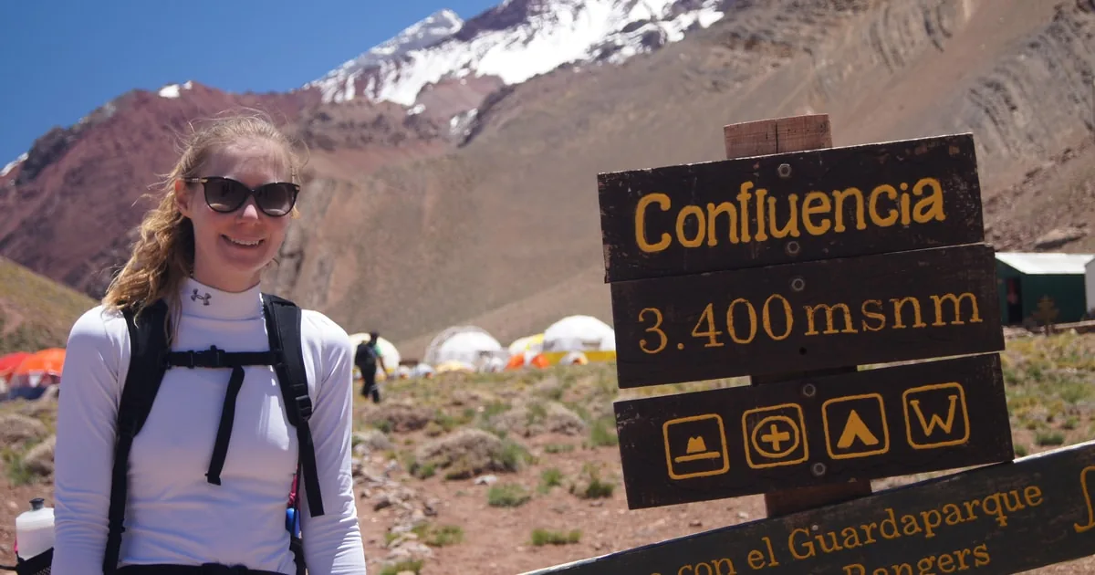 A smiling hiker next to a wooden Confluencia 3,400 m sign, with tents behind