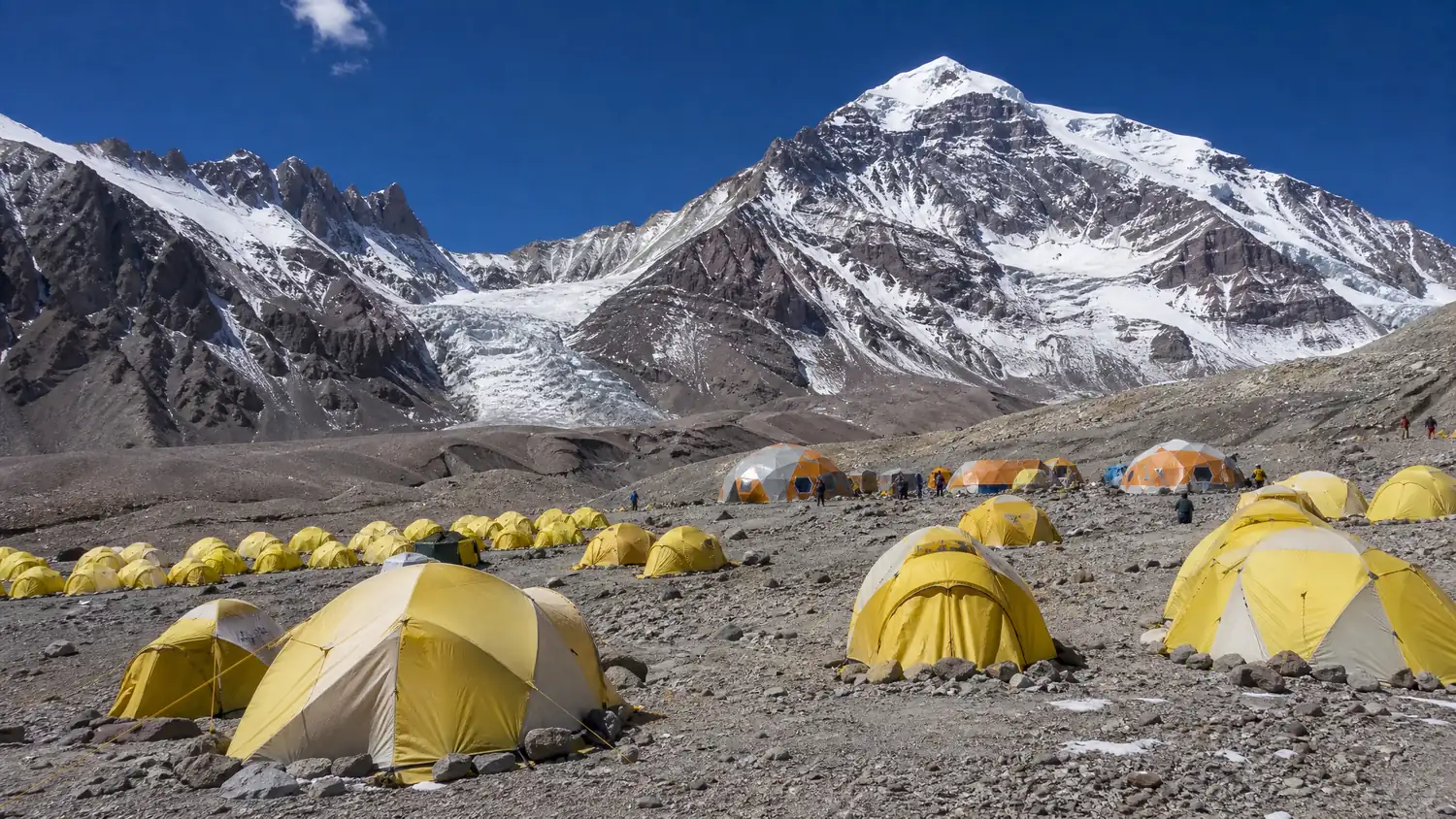 Rows of yellow and orange expedition tents on a rocky moraine at Plaza de Mulas base camp, snowy peaks and a glacier behind