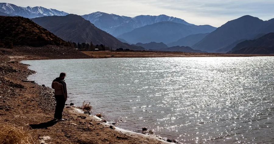 A hiker standing on a stony shore above a sunlit mountain lake in the Andes foothills