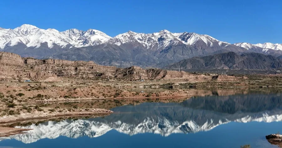 Snow-capped Andes reflected in the still Potrerillos reservoir below reddish cliffs