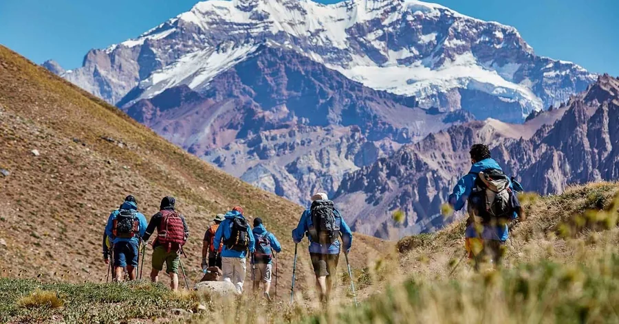 A line of hikers with daypacks walking up a grassy valley towards snowy Aconcagua
